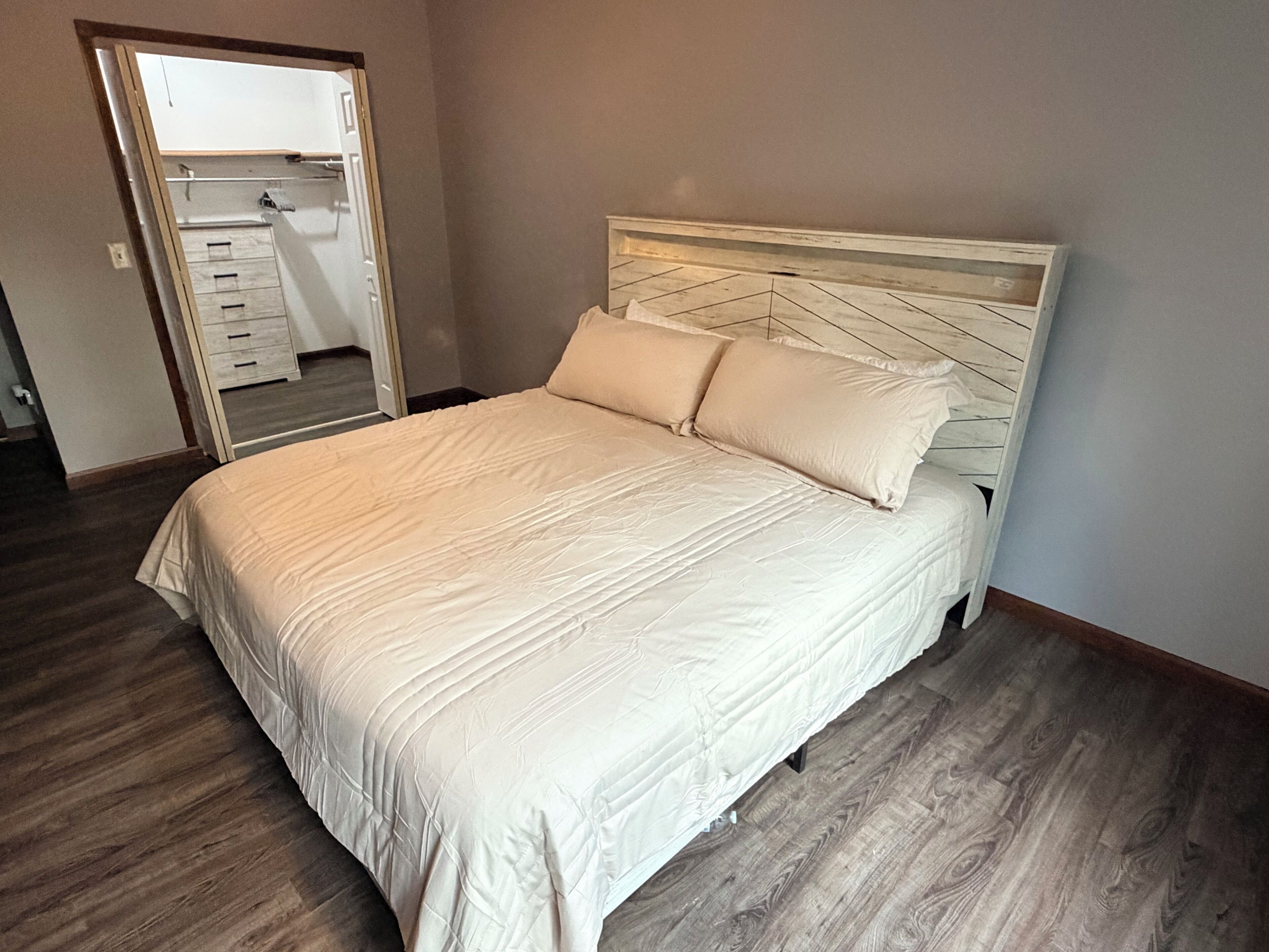 A bedroom with a bed, wooden flooring, and a door leading to another room.