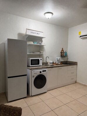 Une petite cuisine équipée d’un réfrigérateur, d’un four à micro-ondes et d’un lave-linge, avec une pancarte indiquant « Réservé aux habitants » et un petit évier.
