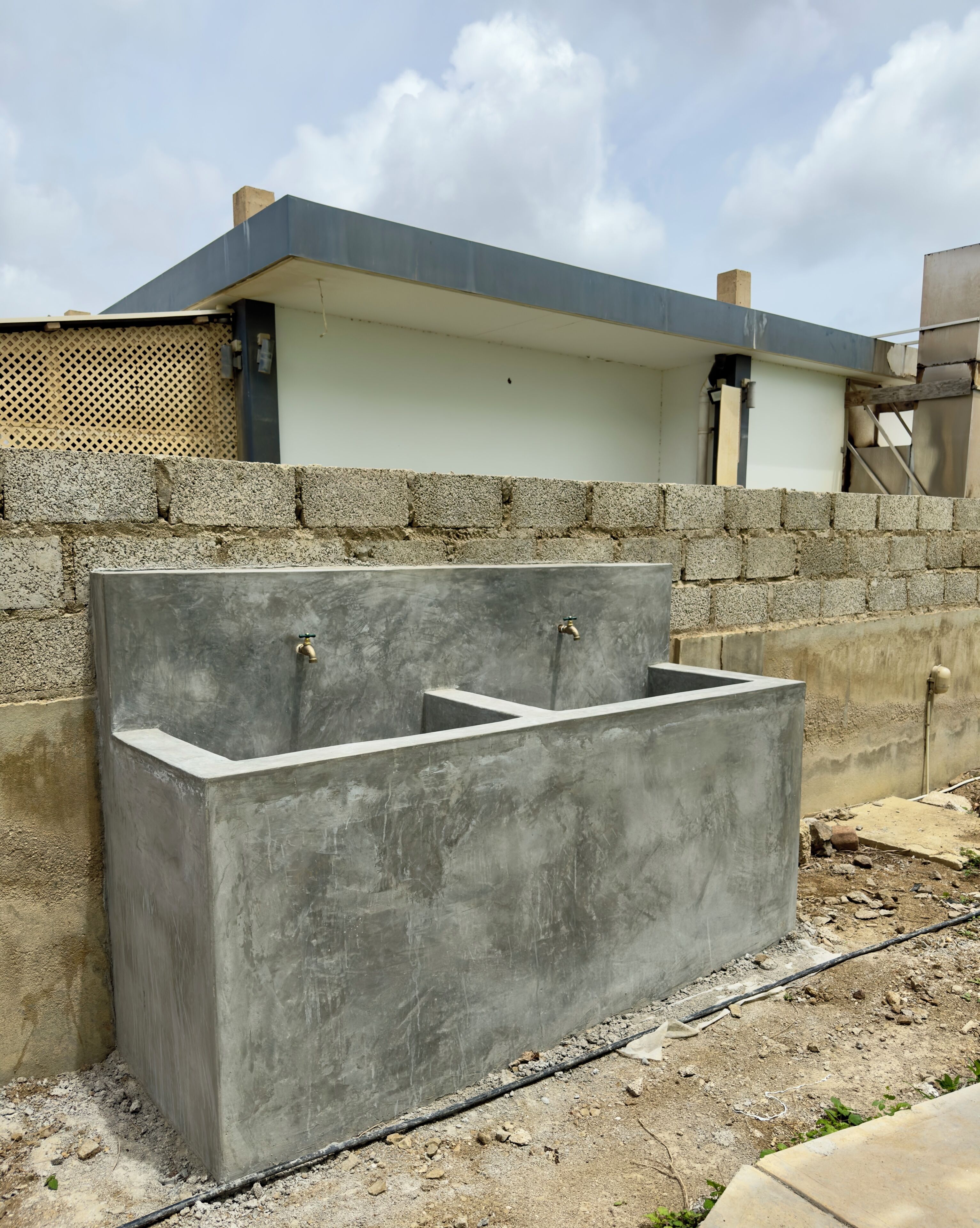 A concrete water feature with two taps, set against a stone wall and a building with a flat roof.