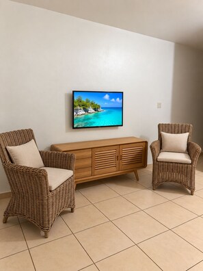 Ein Zimmer mit zwei Korbstühlen, einer Holzkommode und einem Flachbildfernseher, der eine tropische Strandlandschaft zeigt.
