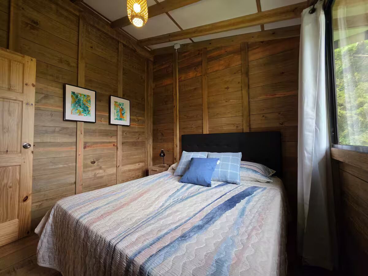 Una habitación de madera con una cama, dos cuadros enmarcados y un ventanal con cortinas.