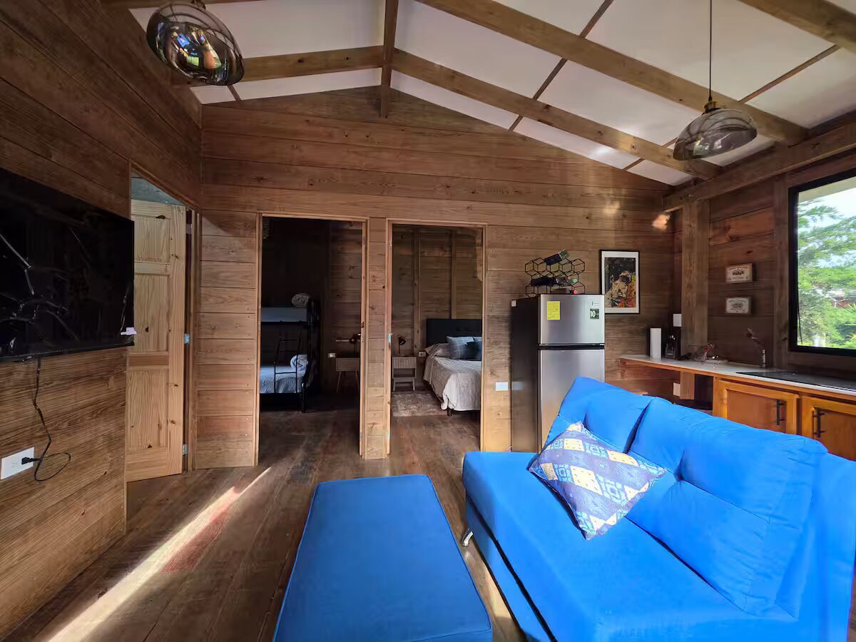Interior acogedor de una cabaña con detalles de madera, un sofá azul, una zona de cocina y un amplio ventanal.