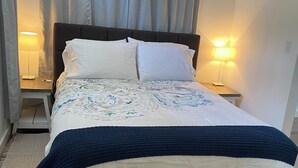 Ein Bett mit dunklem Kopfteil, weißen Kissen und einer gemusterten Tagesdecke. Zwei Nachttische mit Lampen und ein Holzboden sind ebenfalls zu sehen.