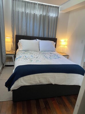 Un letto con una testiera scura, cuscini bianchi e una coperta a motivi. Sono presenti due comodini con lampade e un pavimento in legno.