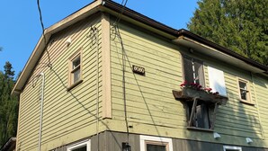 Ein zweistöckiges Haus mit grünen und grauen Fassadenelementen, einer gut sichtbaren Hausnummer '8080' an der Seite und einem kleinen Balkon mit Blumen.