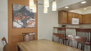 A dining area with a wooden table and chairs, a painting on the wall, and kitchen cabinets above.