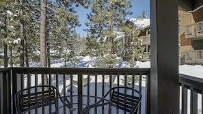 Une terrasse en bois, avec des chaises en métal, donnant sur un paysage enneigé parsemé de pins et sur un bâtiment doté de balcons.