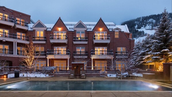 A multi-story hotel with a pool in the foreground, surrounded by snow-covered trees and a mountainous backdrop.