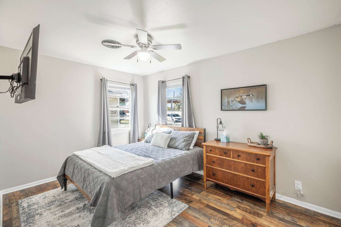 A bedroom with a bed, a wooden dresser, a ceiling fan, and a window with curtains.
