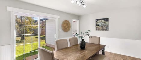 A dining area with a wooden table, beige chairs, a vase with greenery, a ceiling light fixture, and a view through a glass door to an outdoor garden.