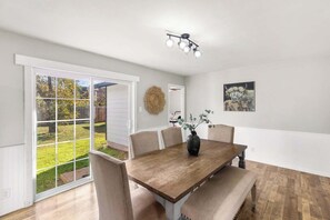 Un coin repas avec une table en bois, des chaises de couleur beige, un vase agrémenté de plantes vertes, un luminaire au plafond et, par une porte vitrée, une vue sur un jardin extérieur.