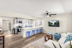 A modern kitchen with white cabinets, a central island with blue bar stools, a dining area with a wooden table, and a living room with a sofa and a mounted TV. - Spacious 4BR with Long Kitchen and Relaxing Patio (Abilene)