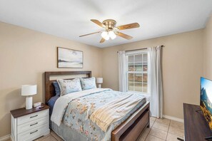 Une chambre à coucher comprenant un lit, des tables de chevet, un ventilateur de plafond, une fenêtre avec des rideaux et un tableau accroché au mur.