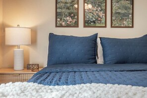 A bedroom with a bed, a nightstand with a lamp and a clock, and three framed paintings on the wall.
