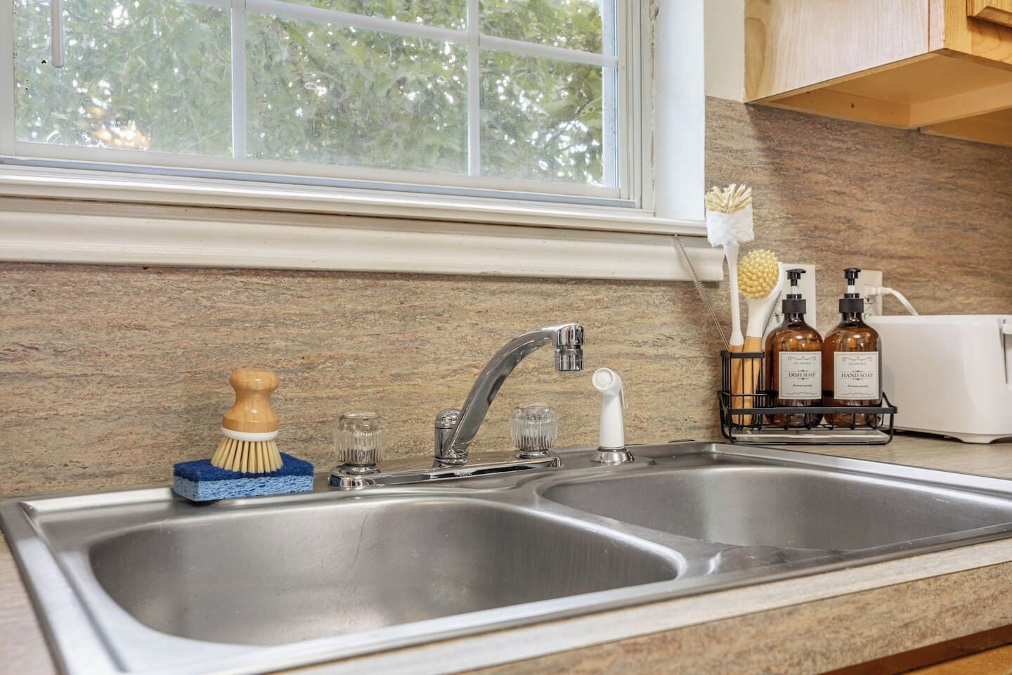 Una cocina con un lavamanos doble de acero inoxidable, una ventana sobre él y una encimera con dispensador de jabón para platos y dispensador de jabón para manos.