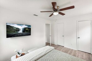 Ein Zimmer mit Deckenventilator, einem Bett mit weißer Matratze und Kissen, einem an der Wand befestigten Fernseher, der einen Wasserfall zeigt, und einer Tür.