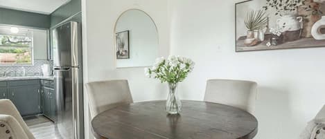 A dining area with a round table, chairs, a vase with flowers, a painting, and a kitchen in the background.