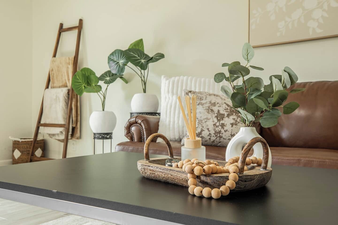 A modern living room with a dark coffee table, a wicker tray with wooden beads, a white vase, and a brown leather couch.
