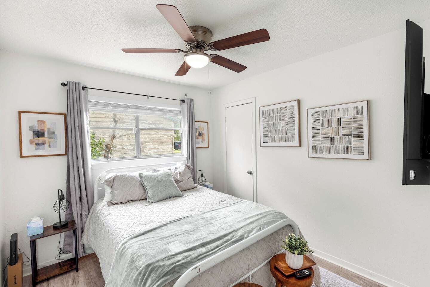 Ein Schlafzimmer mit einem Bett, einem Deckenventilator, einem Fenster mit Vorhängen, einem Nachttisch mit Lampe und gerahmten Kunstwerken an der Wand.