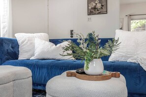 Ein Zimmer mit einem blauen Sofa, weißen Kissen, einer weißen Vase mit Grünpflanzen und einer Fernbedienung auf einem Holzteller.