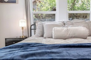 Ein Schlafzimmer mit einem Bett, einem Nachttisch mit Lampe und einem Fenster mit Blick auf Grünflächen.