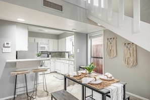 Une cuisine moderne avec des meubles blancs, une table à manger pour quatre personnes et un escalier menant à l’étage supérieur.