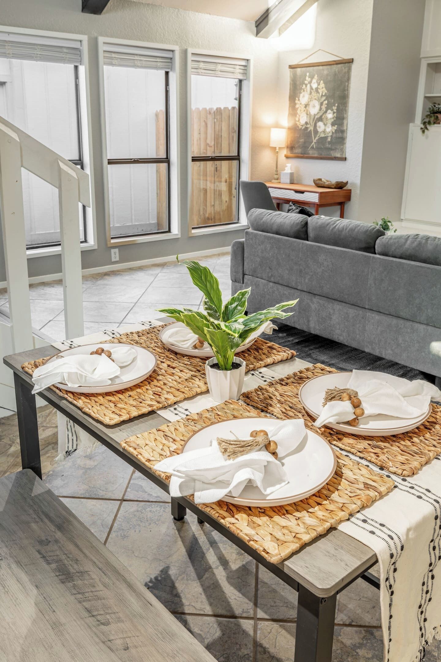 A dining area with a table set for four, a grey sofa, and a large window with blinds.