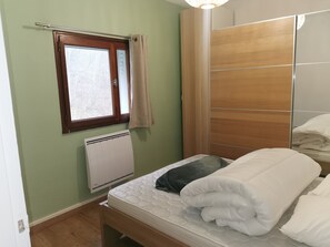 Une chambre avec un lit simple, une fenêtre avec des rideaux, un radiateur et une armoire en bois.