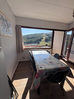 Een kamer met een tafel, stoelen, een raam met uitzicht op een glooiend landschap en een deur naar een buitenruimte.