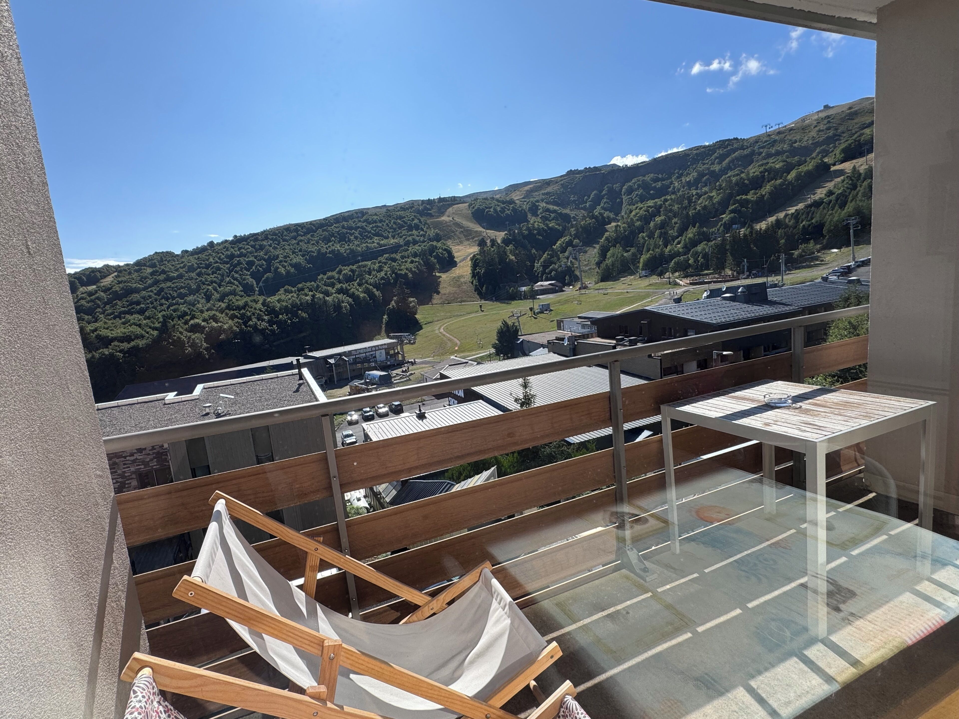 Un balcón con una hamaca, una mesa de vidrio y vistas a un paisaje montañoso con edificios.