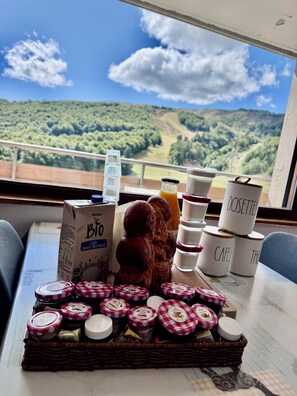 Une table dressée avec des aliments pour le petit-déjeuner, une bouteille de jus d’orange et une vue sur un paysage boisé.