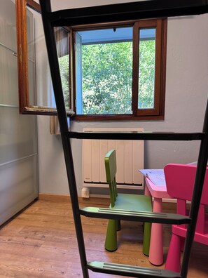 Een kamer met een stapelbed, een raam met uitzicht op bomen, een radiator en een roze stoel.