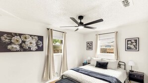 Quarto com cama, ventilador de teto, mesa de cabeceira e janela com cortinas.