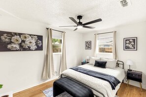 Une chambre à coucher avec un lit, un ventilateur de plafond, une table de chevet et une fenêtre avec des rideaux.