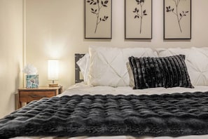 A neatly made bed with a black throw blanket, a bedside table with a lamp and a tissue box, and three framed wall art pieces above the bed.