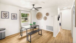Quarto moderno com ventoinha no teto, janela com cortinas, mesa com bancos e obras de arte nas paredes.