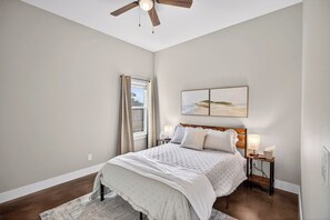 Ein Schlafzimmer mit Bett, Nachttisch, Deckenventilator, Fenster mit Vorhängen und einem Wandbild im Strandhausstil.