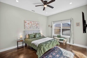 Une chambre à coucher comprenant un lit, des tables de chevet, une fenêtre avec vue, un ventilateur de plafond et un téléviseur fixé au mur.