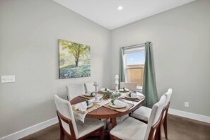 Un coin repas avec une table ronde dressée pour quatre personnes, entourée de chaises, un tableau au mur et une fenêtre avec des rideaux.