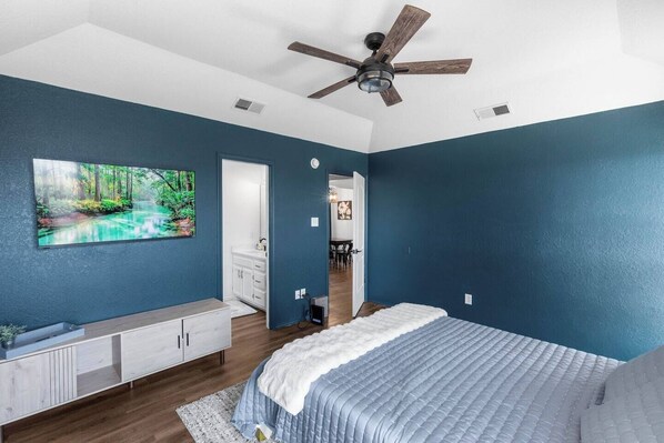 Une chambre à coucher comprenant un lit, un ventilateur de plafond, une commode, un tableau et une porte donnant sur une autre pièce.