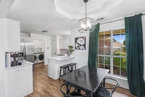 Une cuisine moderne dotée d’un îlot central, d’un coin repas avec une table noire et d’une vue sur une maison en briques, visible à travers de grandes fenêtres.