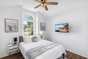 Une chambre à coucher comprenant un lit, un ventilateur de plafond, une fenêtre et deux œuvres d’art accrochées au mur.