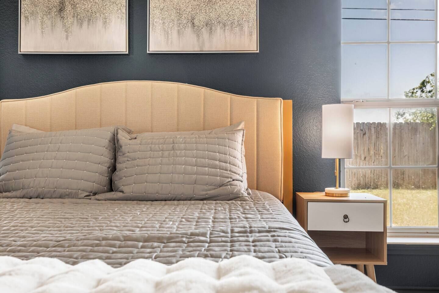 A bedroom with a bed, two pillows, a bedside table with a lamp, and artwork on the wall.