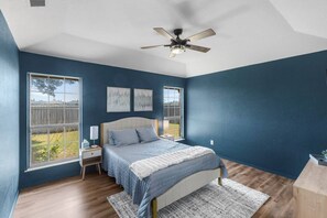 Une chambre à coucher avec un lit, deux tables de chevet, un ventilateur de plafond et de grandes fenêtres.