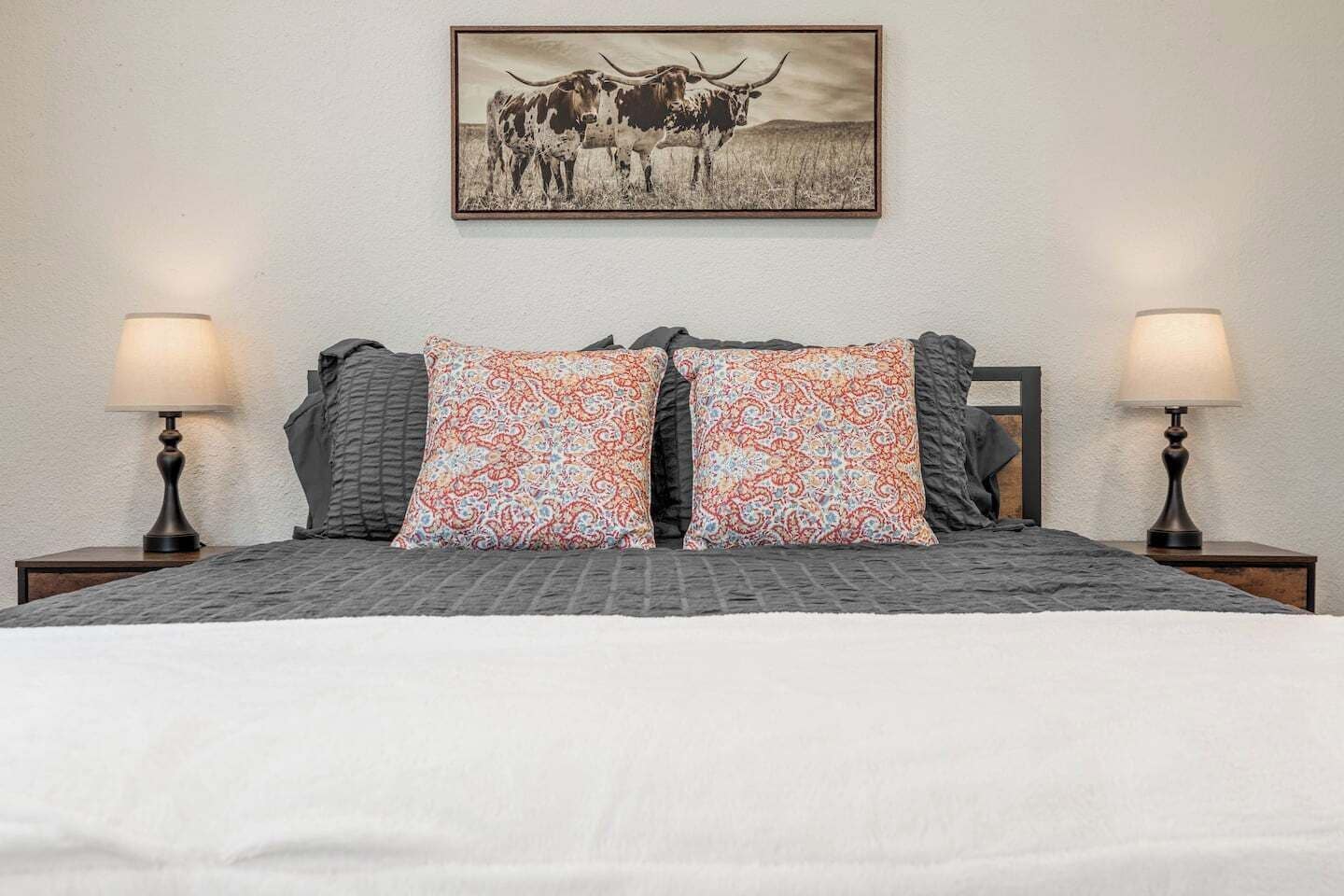 Una cama con colcha estampada, dos almohadas iguales, dos lámparas de noche y un cuadro con vacas en la pared.