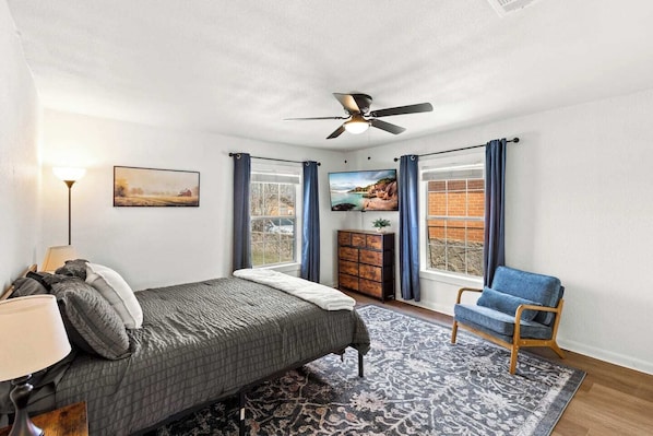 A bedroom with a bed, a chair, a dresser, a ceiling fan, and a window with curtains.