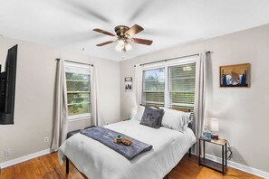 Une chambre à coucher comprenant un lit, un ventilateur de plafond, une fenêtre avec des stores, une table de chevet avec une lampe et un téléviseur fixé au mur.
