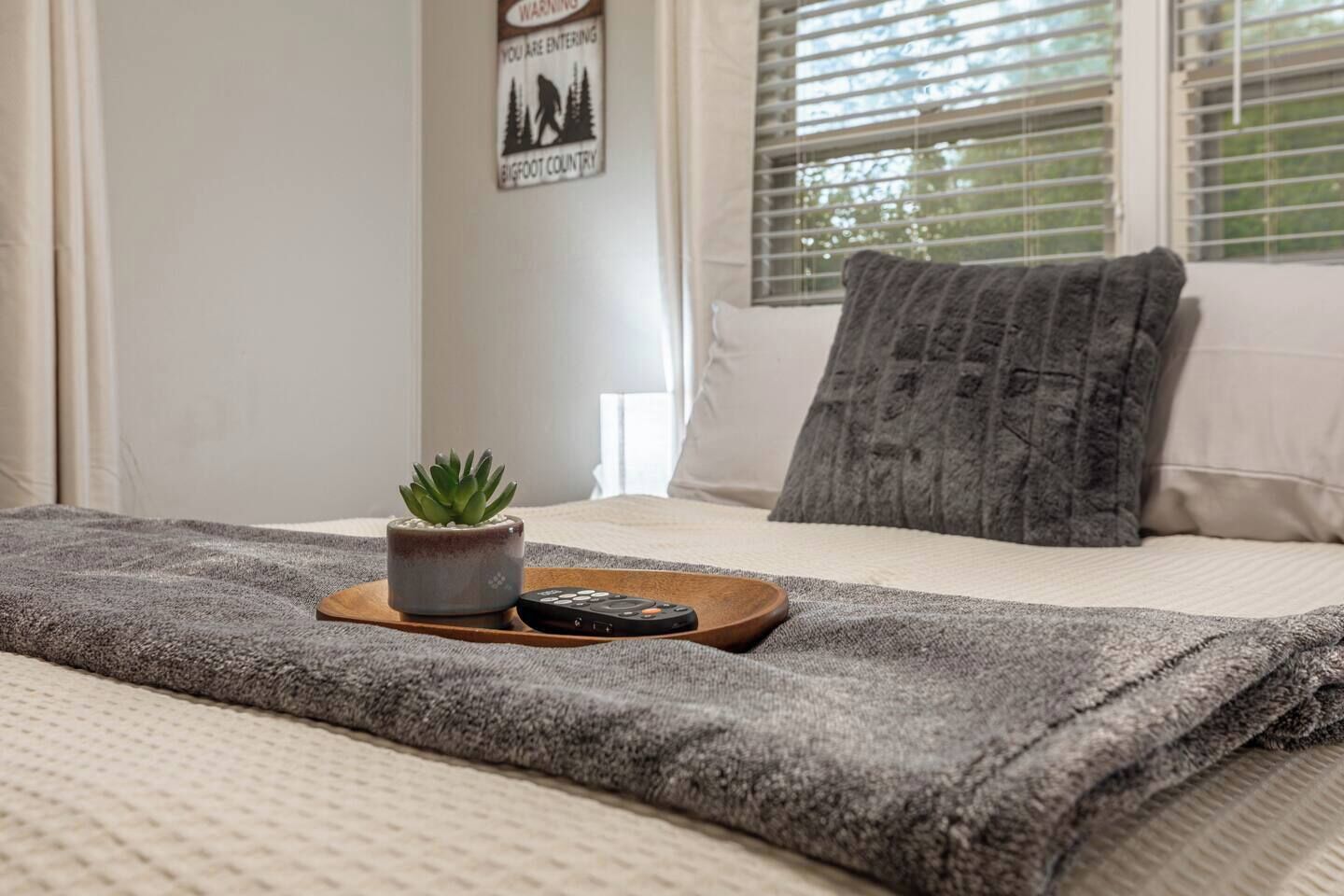 A neatly made bed with a grey blanket, a small potted plant, and a remote control on a wooden tray.