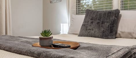 A neatly made bed with a grey blanket, a small potted plant, and a remote control on a wooden tray.