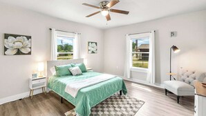 A bedroom with a bed, a ceiling fan, a nightstand, a lamp, a chair, and a window with a view. - Modern 2BR Home with Pool Table & Yard (Abilene)
